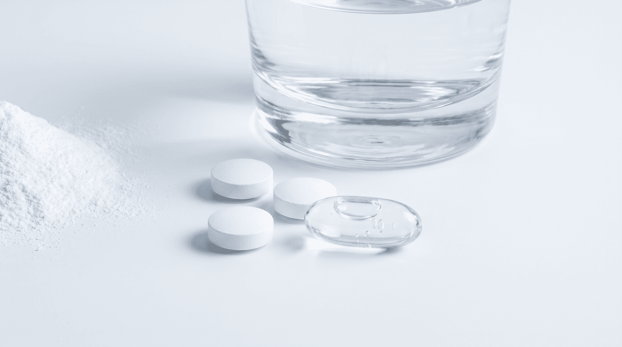 White glutathione tablets beside a liposomal soft-capsule and a glass of water on a clean white surface